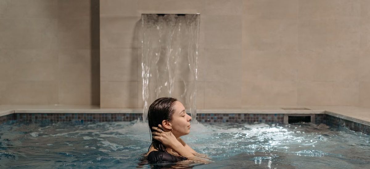 Adult woman relaxing in an indoor swimming pool near a cascading waterfall.